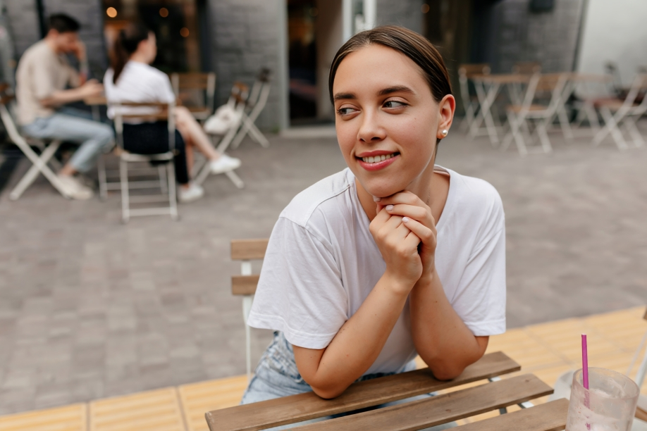 cute lovely woman with happy smile wearing white tshirt is holding hands near face looking aside with happy smile sitting street cafeteria waiting friends