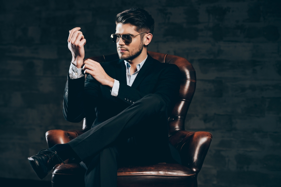 everything must be perfect young handsome man suit sunglasses adjusting sleeve his shirt while sitting leather chair against dark grey background