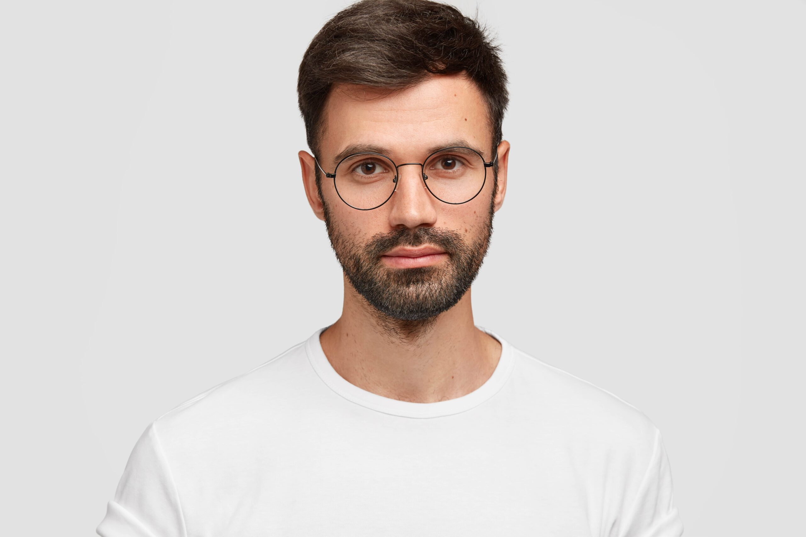headshot handsome male freelancer with appealing look has dark beard mustache looks directly with serious look wears white casual clothes monochrome facial expressions min scaled