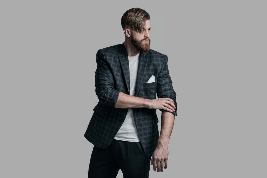 strong confident young hipster looking away adjusting his jacket while standing against grey background